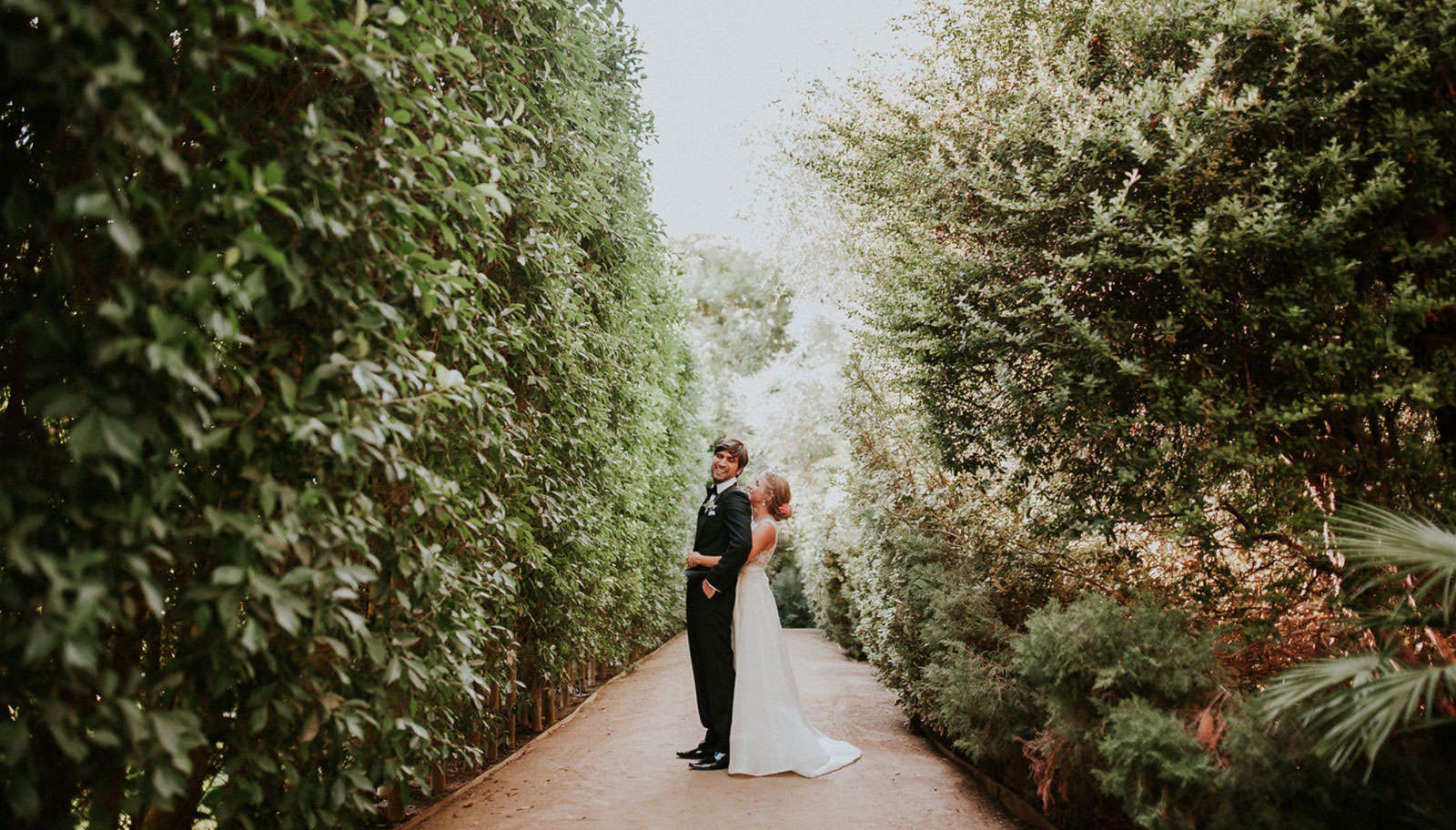 A beautiful couple stands in the center of a vibrant garden path, surrounded by lush greenery and blooming flowers.